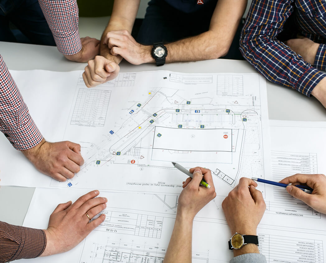 A group of hands pointing and gesturing over detailed architectural plans on a table, engaged in a collaborative discussion.