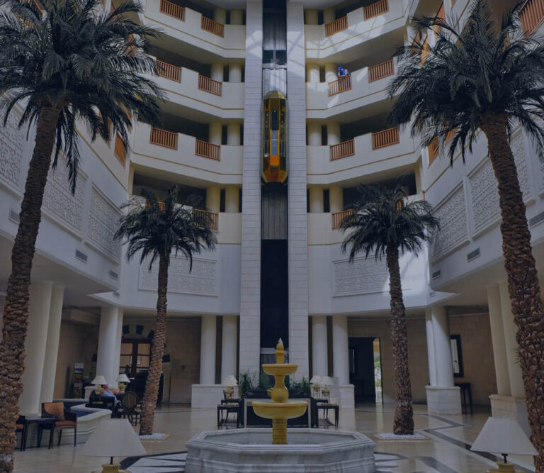 A spacious lobby featuring tall palm trees and a decorative fountain at its center.
