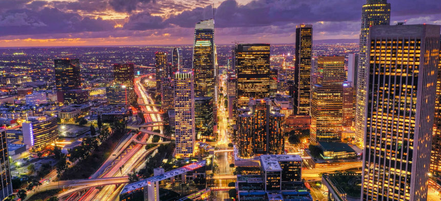 A vibrant city skyline at dusk, showcasing illuminated skyscrapers and busy highways under a colorful sunset sky.
