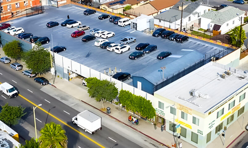 Aerial view of a parking lot filled with cars above a commercial building, alongside a busy street lined with trees.
