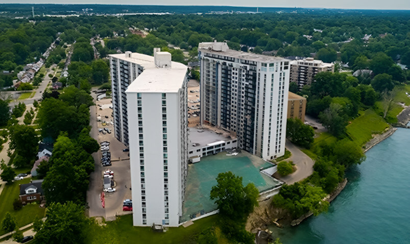 Aerial view of two tall buildings near a lush green landscape and a river, surrounded by trees and nearby homes.