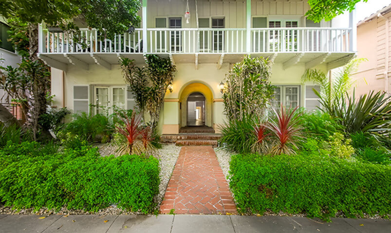 Charming two-story home with a brick walkway, lush greenery, and a welcoming archway entrance framed by vibrant plants.