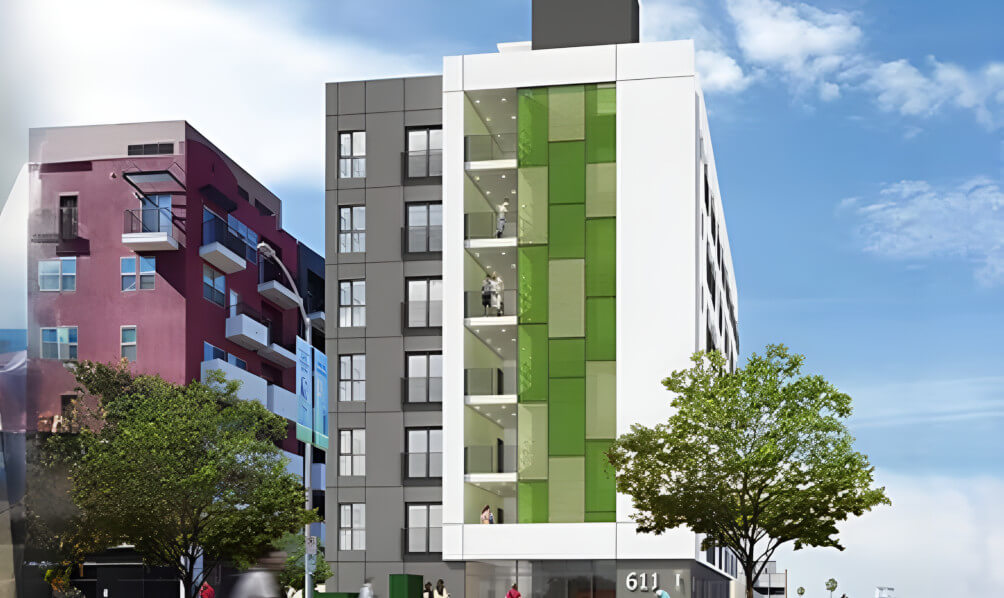 Modern multi-story building featuring green accents, large windows, and balconies, surrounded by trees under a clear blue sky.