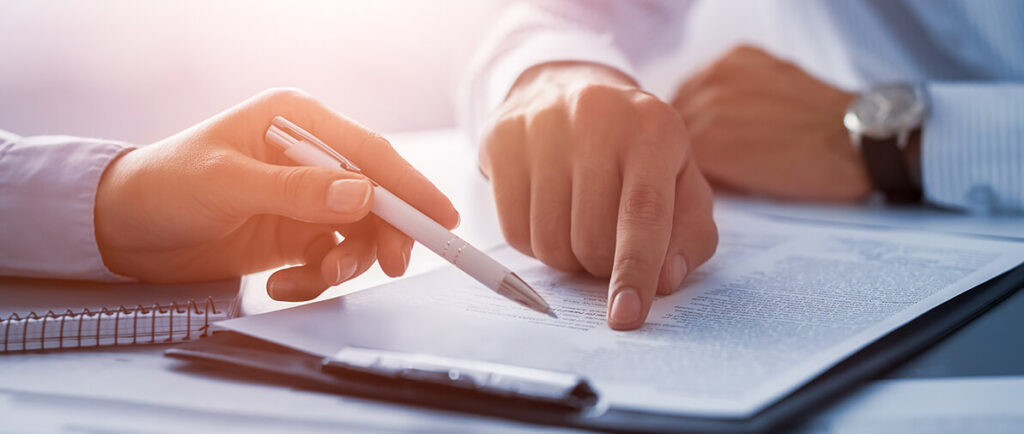 Two hands interact over a document, one holding a pen and the other pointing at the text, in a professional setting.