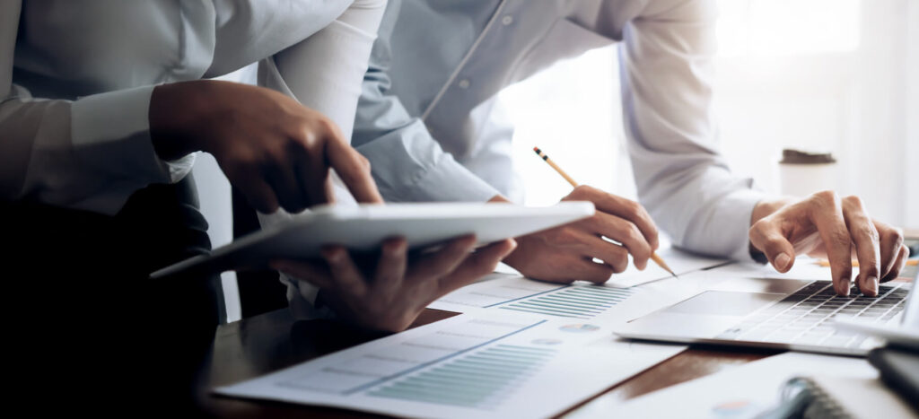 Two professionals analyze documents on a desk, using a tablet and a laptop, with one taking notes with a pencil.