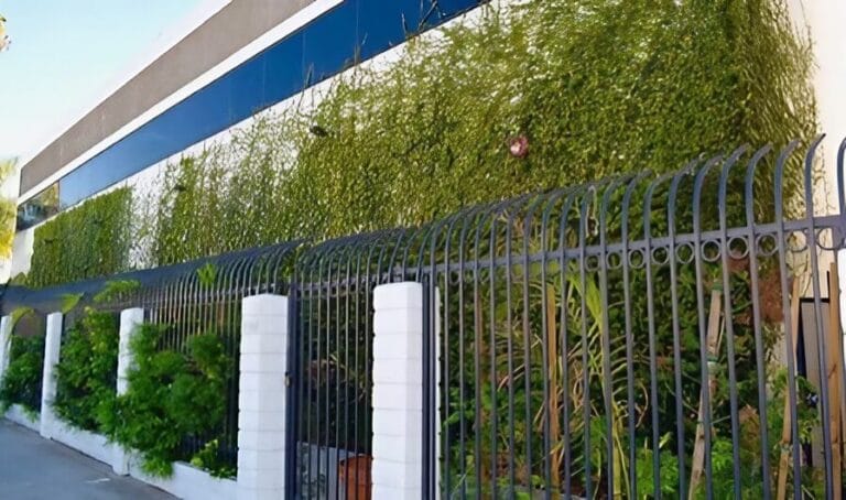 A modern building with a reflective glass facade and a vertical green wall, seen behind a black metal fence with white pillars.