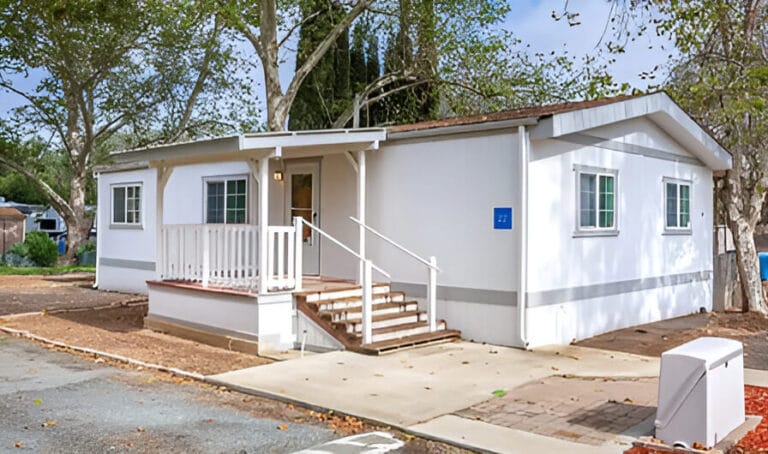 A small white mobile home featuring a porch and stairs, set against a clear sky.