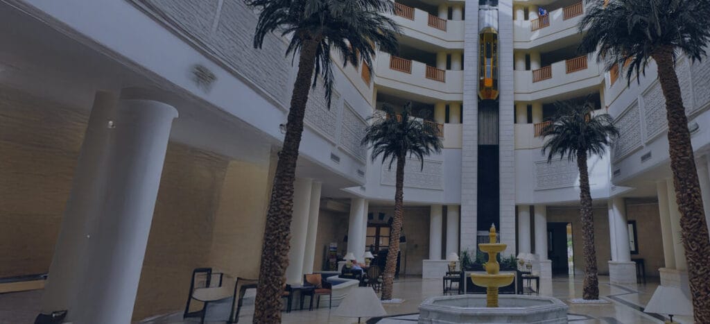 A spacious hotel atrium featuring palm trees, elegant seating areas, and a decorative fountain, exuding a modern yet classic ambiance.
