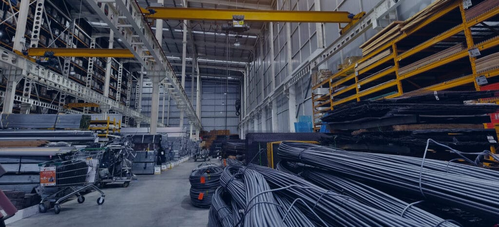 A spacious industrial warehouse filled with stacked metal sheets, rolls of wire, and wooden materials under overhead cranes and shelves.