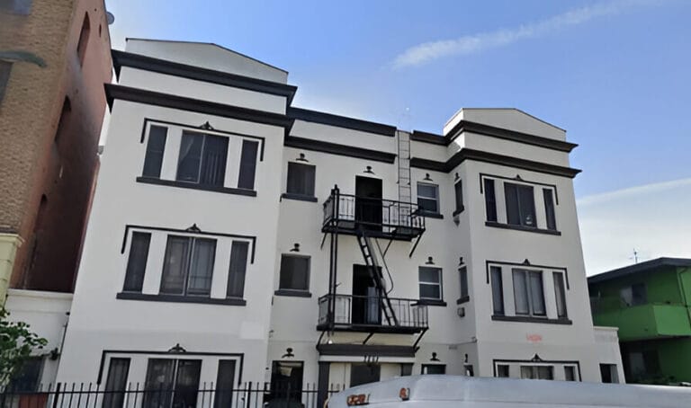 A white apartment building with a white van parked in front, set against a clear blue sky.