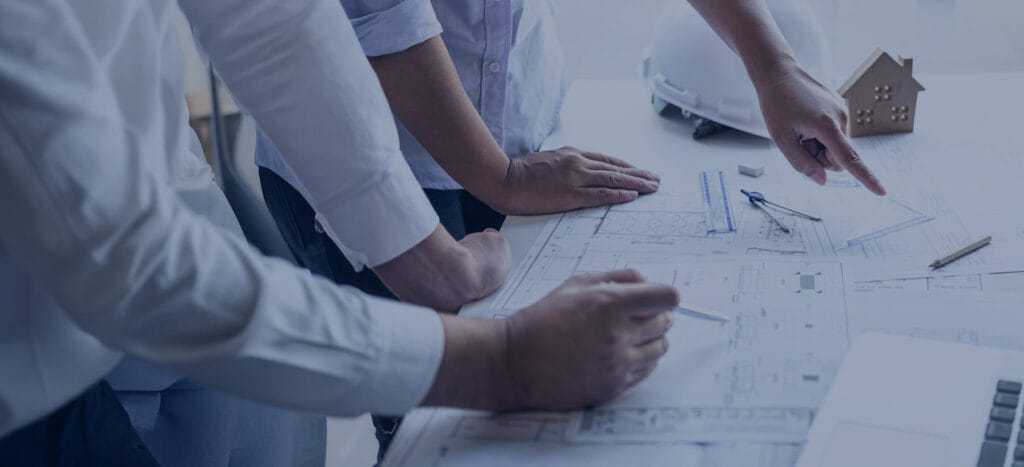 Several people collaborate over architectural blueprints, pointing and writing while tools and a small model house sit nearby.