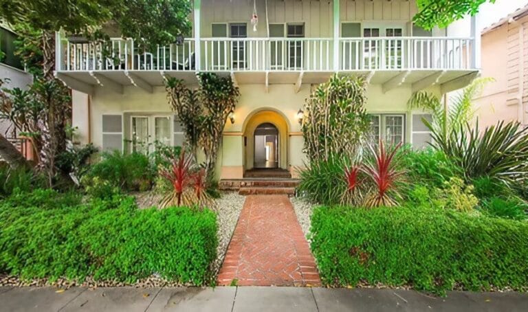 Two-story house with white exterior, arched front entry, balcony, and landscaped garden with green shrubs, red-accented plants, and a brick walkway leading to the door.